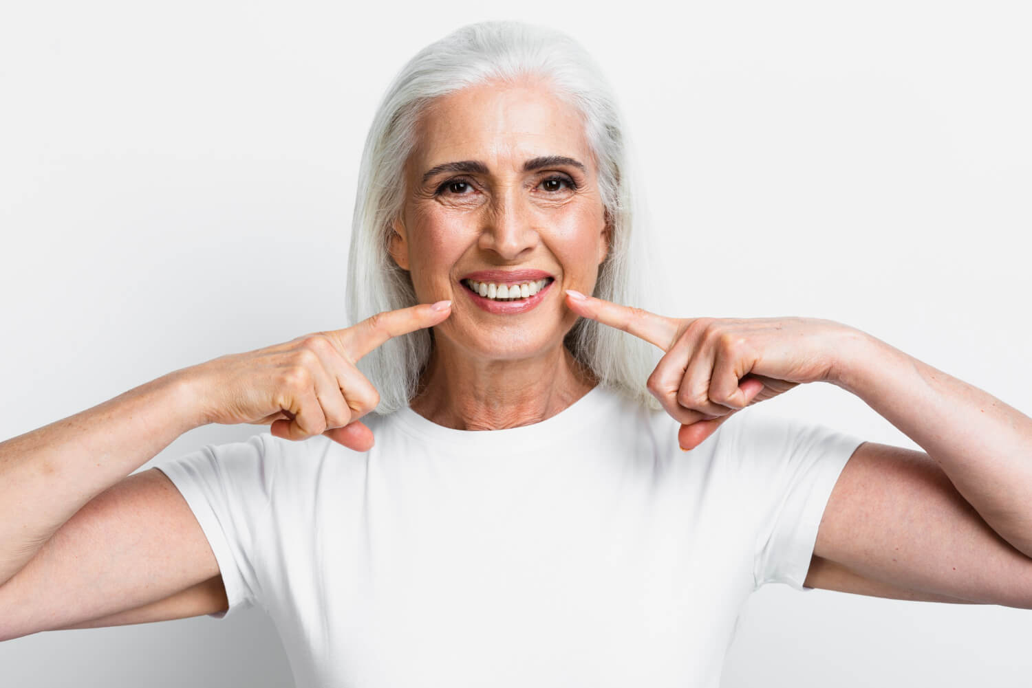 Mulher idosa sorridente aponta para os dentes brancos, representando o resultado de implante dentário em 24 horas na Illuminer em São Bento do Sul.
