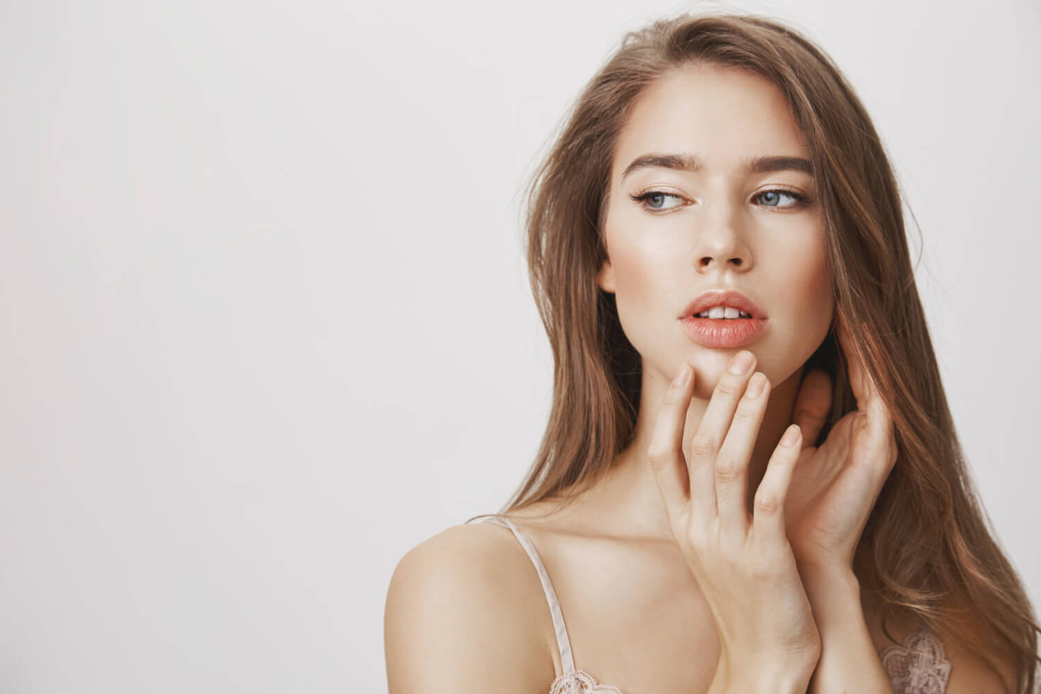 Mulher jovem com preenchimento labial sutil e natural em São Bento do Sul tocando os lábios levemente contra um fundo claro neutro.