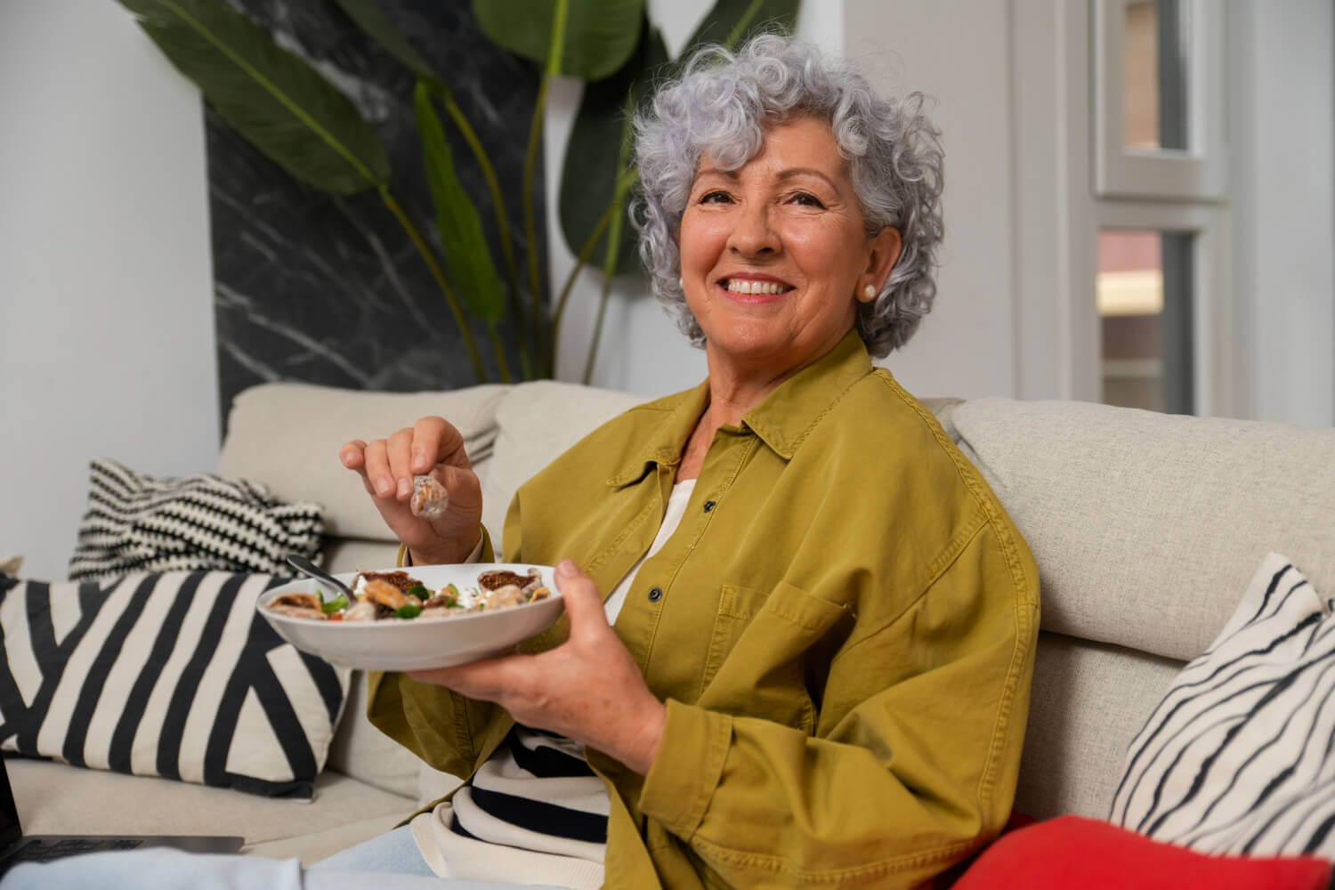 Senhora sorridente no sofá prestes a comer uma refeição, mostrando a segurança na mastigação com prótese protocolo em São Bento do Sul.